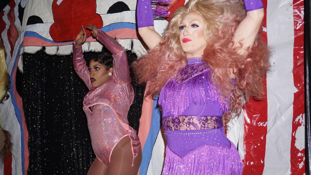 Two drag performers in colorful costumes, one in pink and the other in purple, pose dramatically in front of a vibrant, theatrical backdrop.