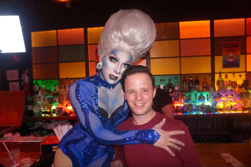 A person in a blue sequined outfit and tall wig strikes a pose with a smiling man at a New York City bar, known for its colorful bottles. It's one of the vibrant spots in town, perfect for catching an unforgettable drag show.