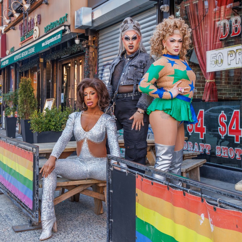 Three drag performers posing on a sidewalk next to a rainbow flag banner outside a bar.