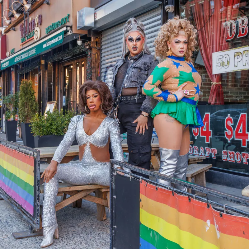 Three drag performers posing on a city street with rainbow flags and festive outfits.
