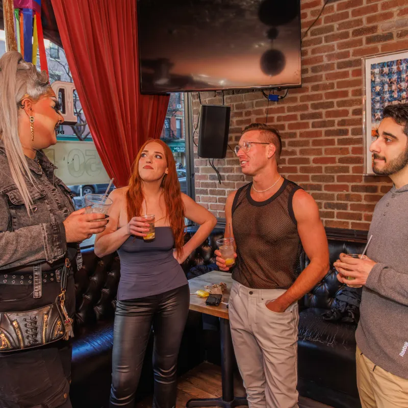 Four people talking and holding drinks in a cozy bar with red curtains and brick walls.