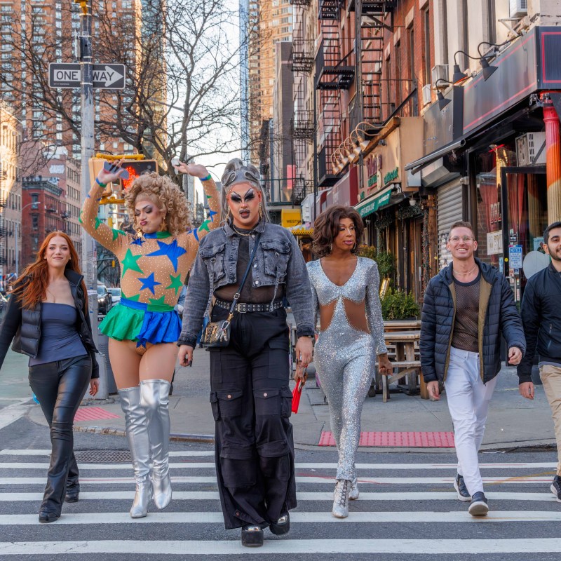 Six diverse people walk on a city crosswalk, dressed in colorful and stylish outfits.