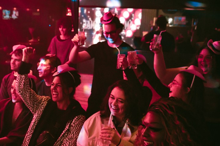 Group of people in a nightclub celebrating, wearing hats and holding drinks, under red lighting.