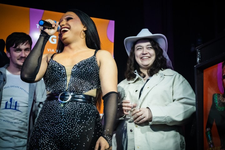 Person in black sequin outfit singing, person in white hat smiling, with a microphone and drink on stage.