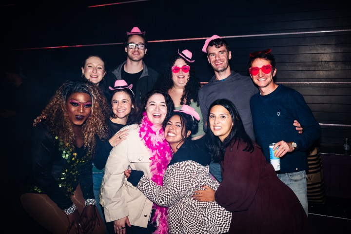 Group of 10 people at a party, some wearing pink hats and sunglasses, posing happily with a performer in costume.