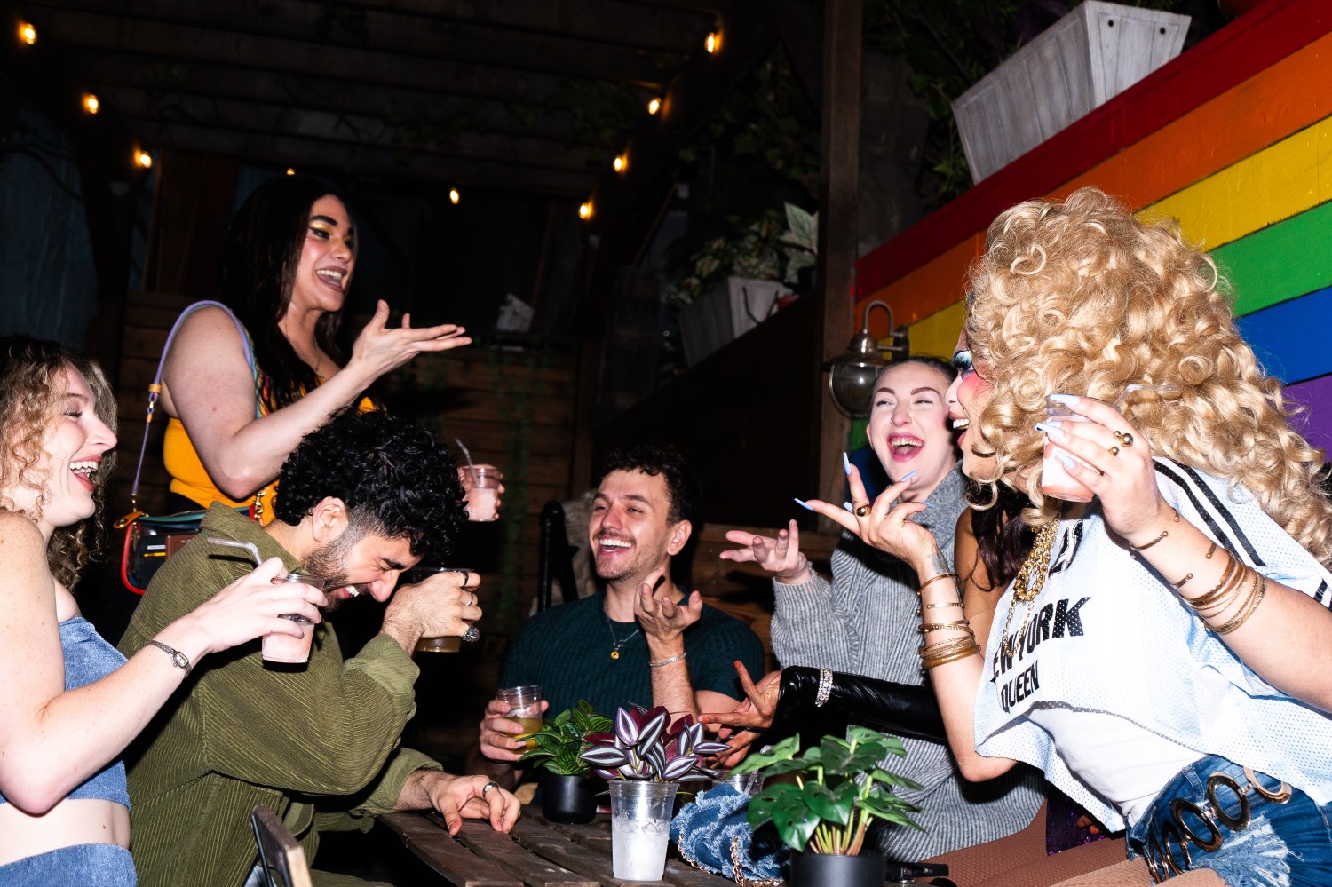 Group of friends laughing and talking at a table with drinks, indoor setting with rainbow background.