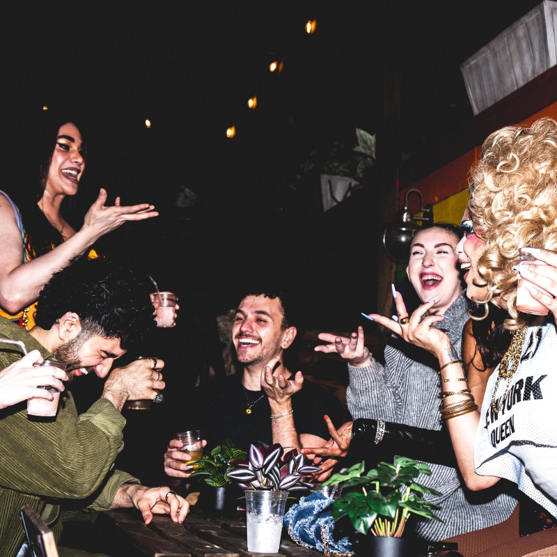 Group of people laughing and celebrating at an outdoor party with drinks and a rainbow wall.