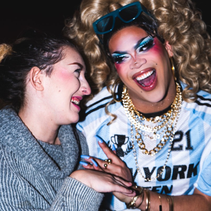 Two people laughing, one in drag with bold makeup and blonde wig, wearing a sporty top.