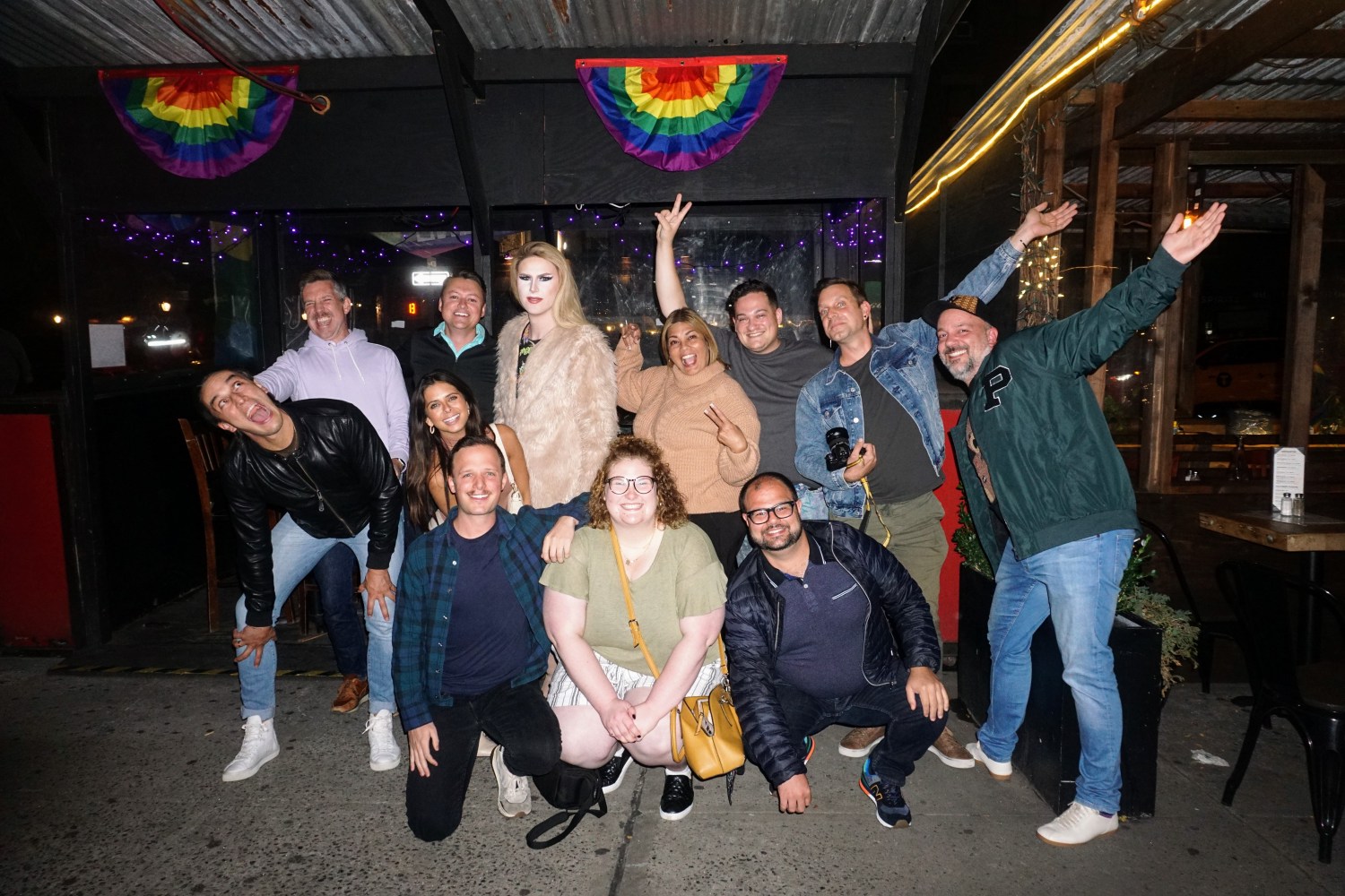 Group of people posing happily on a sidewalk under rainbow flags.