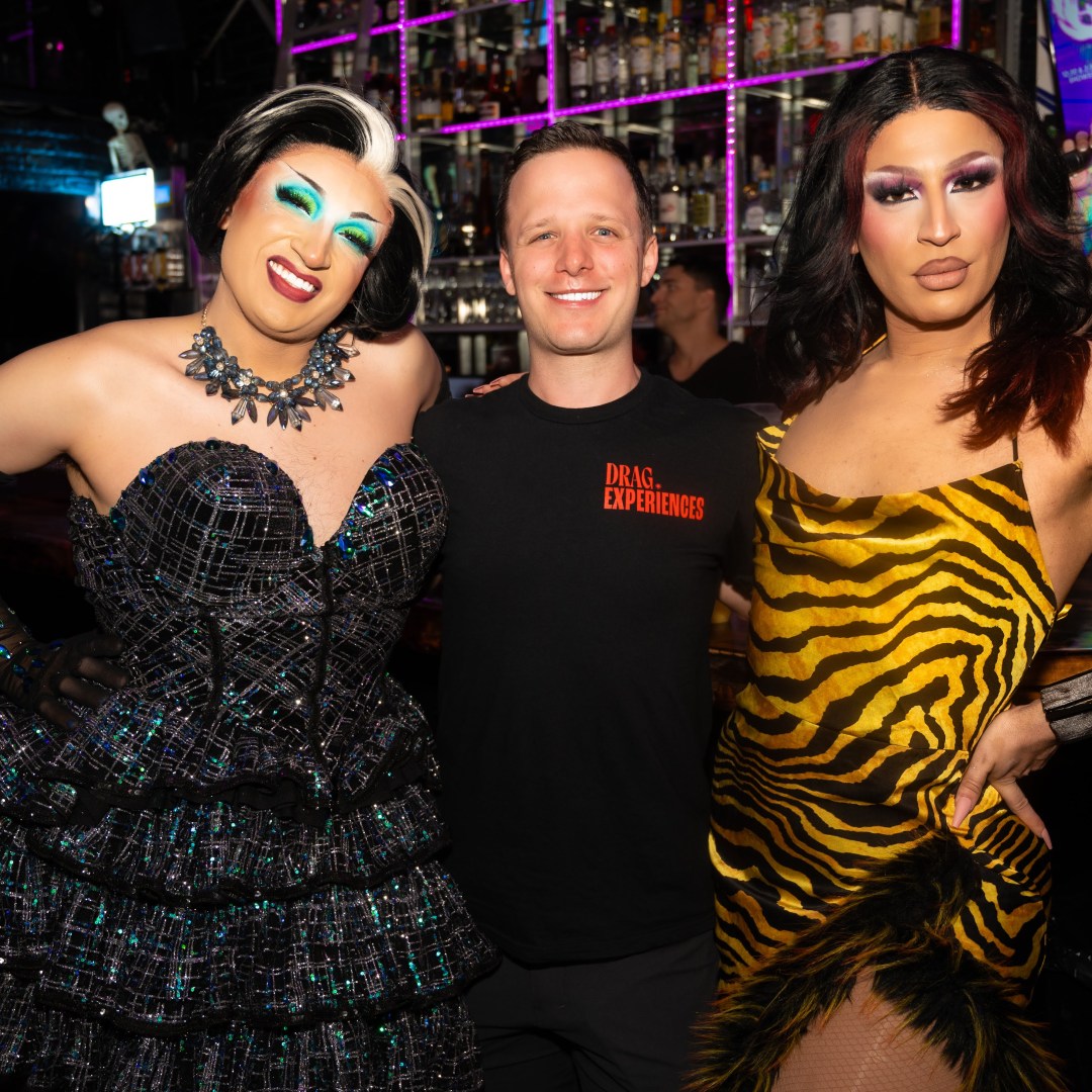 Three people posing in a bar, two wearing colorful drag outfits, with a neon-lit bar in the background.