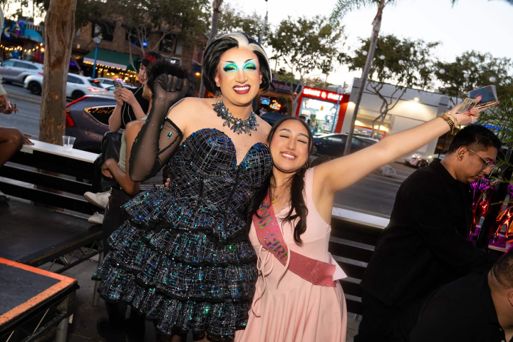 Two people posing and smiling outdoors, one in a sparkly dress, the other with a sash, surrounded by others.