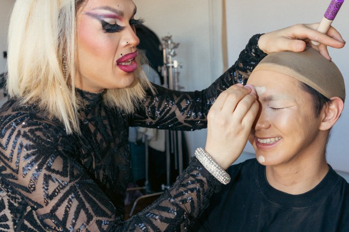Person with makeup applies concealer on another smiling person's forehead in a studio setting.