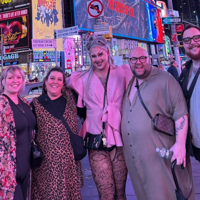 Five people smiling in Times Square at night with bright billboards in the background.