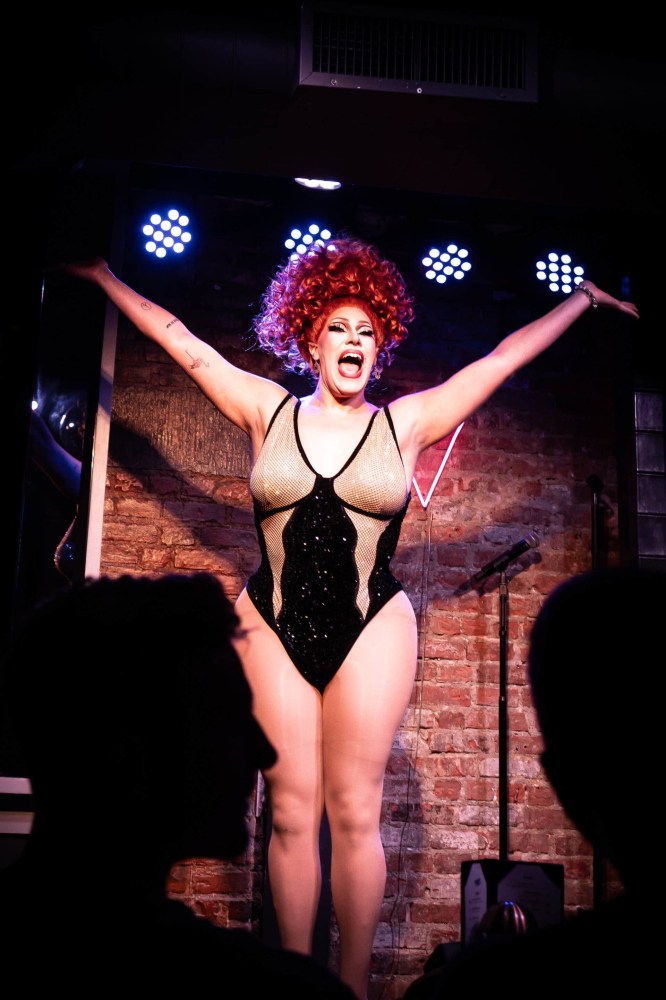 Drag performer in sparkly bodysuit on stage, arms outstretched, with audience silhouettes in foreground.