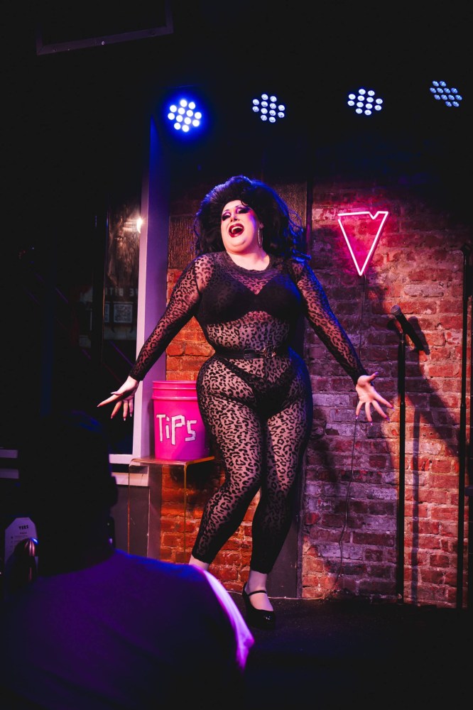 Drag performer in leopard outfit on stage with tip bucket and neon sign.