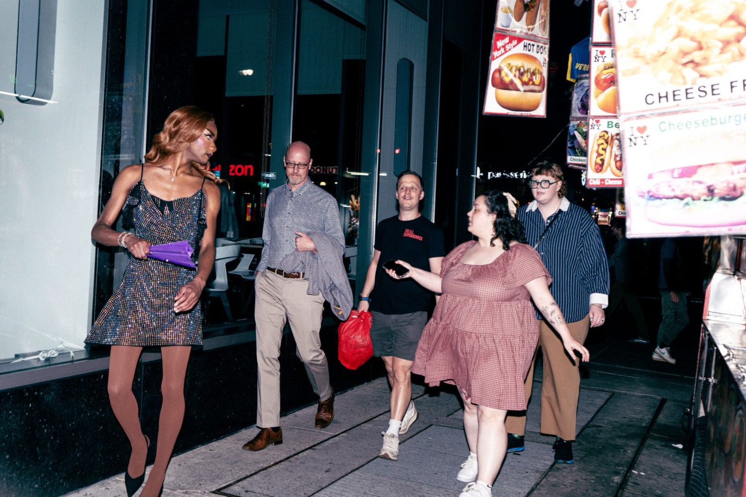 People walking on a city street at night near a restaurant with food signs.
